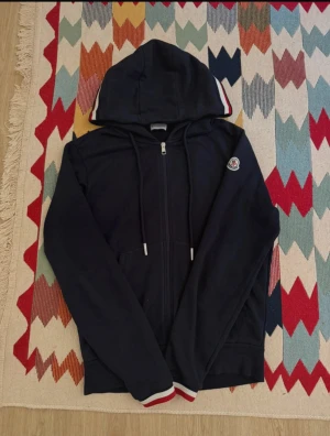 Moncler zip up hoodie Mörkblå  - Säljer denna mörkblå zip-hoodie i väldigt fint skick. Hoodien är perfekt för dig som vill ha ett plagg som både är bekvämt och stilrent, och som funkar i stort sett året runt. Färgen är en klassisk mörkblå ton som passar till allt och ger ett rent, fräscht intryck. På ena ärmen sitter en patch som ger plagget en lite mer exklusiv känsla. Den har även snygga tricolor-detaljer på huvan och vid muddar, vilket gör att den sticker ut på ett diskret men stiligt sätt.  