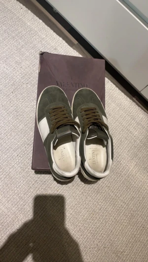 Valentino Garavani Flycrew  - Snygga Valentino Garavani sneakers i olivgrönt mocka med vita skinnpaneler och bruna snören. Klassisk rund tå och platt sula med ikoniska nitar på hälen. Kommer med originalkartong och extra skosnören. Perfekt för dig som gillar exklusiva streetwear-vibbar. Byter endast! Kläder & Skor inkl jackor e najs