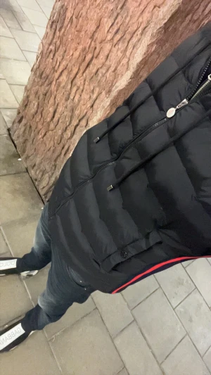 Moncler jacka - Svart cardigan från Moncler med quiltad framsida, dragkedja och hög krage. Sidorna har mörkblå och röda ränder samt Moncler-logga baktill. Västen har stickade partier på axlar och rygg för extra komfort och stil.