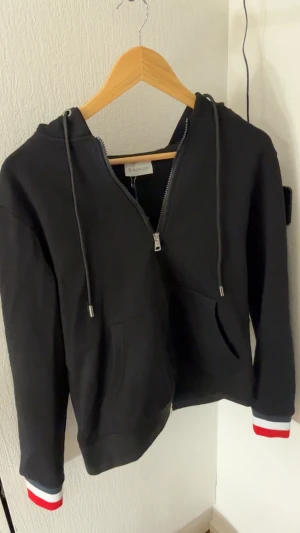 Svart Moncler hoodie med dragkedja - Svart hoodie från Moncler med hel dragkedja, huva och snörning. Tröjan har Moncler-logga på ärmen och coola ränder i rött, vitt och blått på muddar och huva. Tillverkad i mjuk bomull, perfekt för chill dagar. Snygg och sportig vibe!