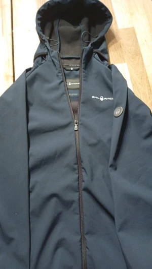 Mörkblå vindjacka från Sail Racing - Snygg mörkblå vindjacka från Sail Racing med huva och dragkedja framtill. Jackan har logotyp på bröstet och ärmen, samt en metallbadge med Sail Racing-symbolen. Materialet är slätt och känns som polyester, perfekt för blåsiga dagar.