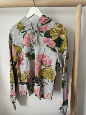 Grå blommig hoodie från H&M - Grå hoodie från H&M med stort blommönster i rosa och grönt. Tröjan har huva med snörning och långa ärmar. Mjuk och skön bomullsblandning.