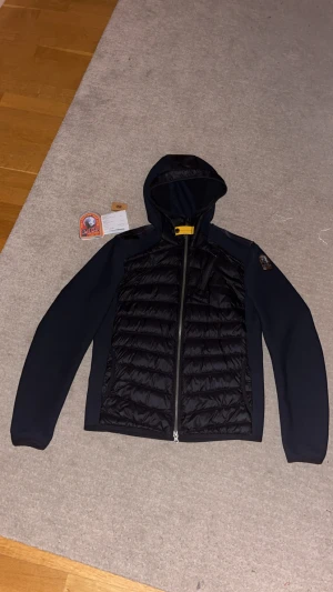 Svart pufferjacka från Parajumpers XS - Svart pufferjacka från Parajumpers i storlek XS. Jackan har huva, quiltad framsida och dragkedja hela vägen. Materialmix med polyester och syntet, samt snygga detaljer som gul rem vid halsen och märkets patch på ärmen. Perfekt för kyliga dagar.