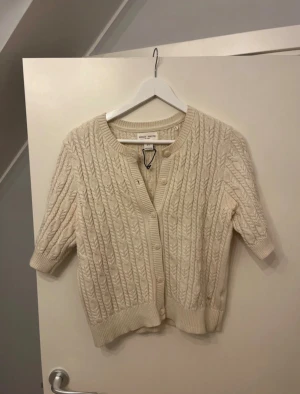 Beige stickad kofta Holly & Whyte - Superfin beige stickad kofta från Holly & Whyte med kort ärm och snyggt kabelstickat mönster. Rund halsringning, knappar framtill och ribbade muddar. Perfekt att slänga över en topp för en chill look.