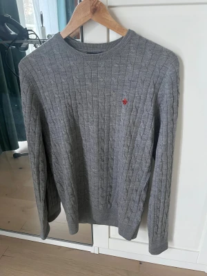 Grå kabelstickad tröja Morris M. Reserverad! - Snygg grå kabelstickad tröja från Morris i 100% merinoull. Rund hals, långärmad och med röd broderad logga på bröstet. Perfekt för dig som gillar klassisk stil med modern touch. Passar dig som vill ha en normal passform och mjukt material.