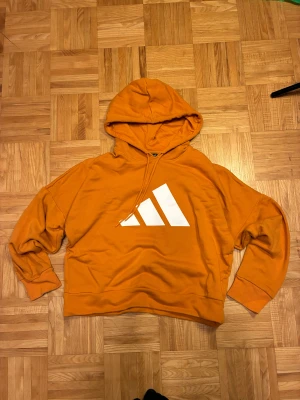Orange hoodie från Adidas med logga - Snygg orange hoodie från Adidas med stor vit logga på bröstet och snörning vid huvan. Tröjan har lång ärm, ribbade muddar och är tillverkad i mjuk bomull. Perfekt för dig som gillar sportig och avslappnad stil.