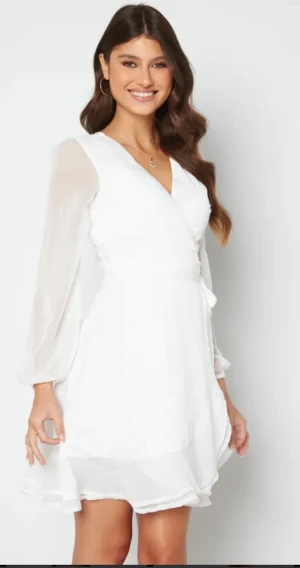 Vit omlottklänning från Bubbelroom - Supersöt vit omlottklänning från Bubbelroom med v-ringning och långa, transparenta ärmar. Klänningen har volangdetaljer nedtill och ett knytband i midjan som ger en snygg siluett. Perfekt för sommaren och studenten!