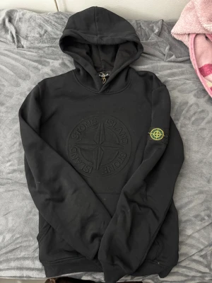 Svart hoodie från Stone Island - Svart hoodie från Stone Island med stort broderat logomärke på bröstet och klassisk patch på vänster ärm. Tröjan har huva och ribbade muddar vid ärmslut och nederkant. Tillverkad i mjuk bomull, perfekt för chill dagar.