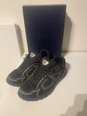 B30  - Säljer ett par svarta sneakers med snygga grå detaljer och meshpaneler. Skorna har rund tå, snörning och en chunky platt sula. Märket syns tydligt på plösen och boxen ingår. Perfekt för dig som gillar exklusiva och trendiga sneakers.