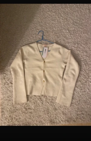 Beige kofta från Gina Tricot - Snygg beige kofta från Gina Tricot med v-ringning och tre guldfärgade knappar framtill. Koftan är stickad och har långa ärmar, perfekt för lager-på-lager. Materialet är 50% LENZING ECOVERO viskos för en mjuk och skön känsla.