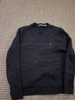 Mörkblå stickad tröja Polo Ralph Lauren - Snygg mörkblå stickad tröja från Polo Ralph Lauren med klassiskt kabelstickat mönster och röd broderad logga på bröstet. Rund hals och ribbade muddar vid ärmslut och nederkant. Perfekt för dig som gillar stilren och tidlös design.
