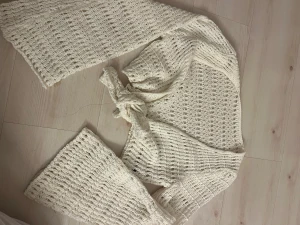 Vit stickad bolerotopp med knyt - Säljer en vit bolerotopp i stickat material med grov virkning och långa ärmar. Toppen har en snygg knytning framtill och är perfekt för att ge din outfit en chill och bohemisk vibe. Passar dig som gillar unika detaljer och vill ha något som sticker ut.