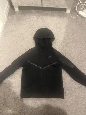 Svart Nike hoodie med dragkedja - Svart hoodie från Nike med hel dragkedja och huva. Snygga svarta detaljer i form av ett diagonalt band över bröstet och en ficka med dragkedja på ärmen. Tröjan är gjord i mjuk bomull och har långärmad passform. Perfekt för dig som gillar sportig och clean stil.