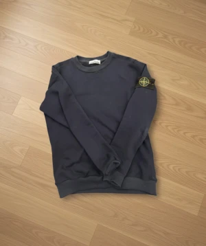 Mörkblå Stone Island sweatshirt - Snygg mörkblå sweatshirt från Stone Island med klassisk patch på vänster ärm. Tröjan har rund hals, ribbade muddar och är tillverkad i mjuk bomull. Perfekt för dig som gillar streetwear och vill ha en clean look med ikonisk detalj.