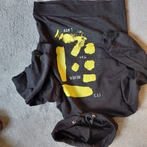 Svart hoodie med gul print från NA-KD - Cool svart hoodie från NA-KD i storlek S med huva och snörning. Framsidan har ett grafiskt tryck i gult med texten 'IT'S NEVER TOO LATE'. Tröjan är tillverkad i mjuk bomull och har långa ärmar samt ribbade muddar vid ärmslut och nederkant.