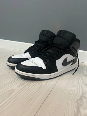 Nike Air Jordan 1 svart/vit sneakers - Nike Air Jordan 1 sneakers i svart och vitt med grå detaljer. Klassisk high-top modell med snörning och rund tå. Swoosh-logga på sidan och Air Jordan-märke vid ankeln. Tillverkade i skinn med perforerad tå för extra ventilation. Perfekt för dig som gillar streetwear och ikoniska sneakers. Helt äkta köpta från Nikes hemsida och är i nytt skick