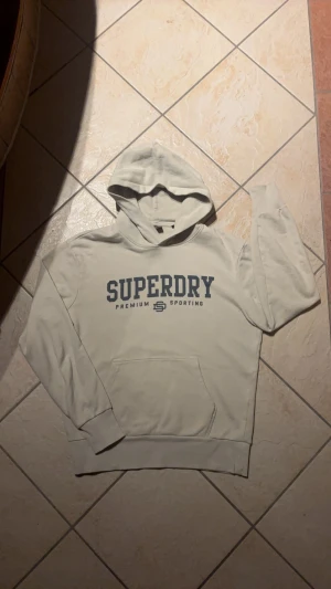 Beige hoodie från Superdry i M - Snygg beige hoodie från Superdry med stor mörkblå logga på bröstet. Tröjan har huva, lång ärm och en klassisk känguruficka framtill. Mjuk bomullskvalitet som är skön att ha på sig. Perfekt för chill eller när du vill ha en clean streetstyle.