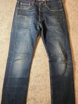 Levi's 501 blå jeans W28 L32 - Klassiska Levi's 501 jeans i blå denim med raka ben och normal passform. Snyggt slitna detaljer och femficksmodell. Tidlös design med knappgylf och ikonisk läderpatch bak i midjan. Perfekta för dig som gillar en autentisk jeanslook.