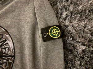 Grå sweatshirt från Stone Island - Säljer en grå sweatshirt från Stone Island med stort svart logomärke i materialmix på bröstet och klassisk patch på ärmen. Tröjan har rund halsringning och långa ärmar med ribbade muddar. Perfekt för dig som gillar streetwear och vill sticka ut.