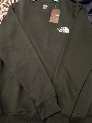 Svart sweatshirt från The North Face - Svart sweatshirt från The North Face med klassisk logga i vitt på bröstet. Tröjan har rund hals, långa ärmar och ribbade muddar vid ärmslut och nederkant. Perfekt för dig som gillar enkel och stilren streetwear.