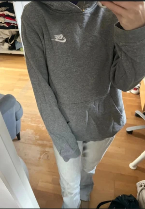 Grå hoodie från Nike med huva - Säljer en grå hoodie från Nike med klassisk logga på bröstet. Tröjan har huva, stor magficka och långa ärmar. Perfekt för chill dagar och enkel att matcha med jeans eller mjukis. Materialet är mjuk bomull och polyesterblandning för skön komfort.