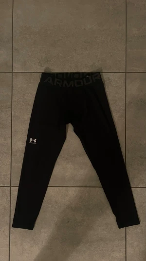 Svarta tights från Under Armour - Svarta tights från Under Armour med logga på benet och bred resår i midjan med text. Sitter tight och är perfekta för träning. Gjorda i stretchigt syntetmaterial som andas och ger bra rörelsefrihet.