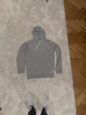Grå hoodie från H&M med huva - En stilren grå hoodie från H&M i regular fit, storlek S. Tröjan har en klassisk huva med vita snören och en stor magficka framtill. Mjuk bomullsmix som är perfekt för chill dagar. Långärmad och enkel att matcha med jeans eller joggers.
