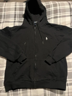 Svart hoodie från Polo Ralph Lauren - Svart hoodie från Polo Ralph Lauren med klassisk vit broderad logga på bröstet. Tröjan har huva med snörning, dragkedja framtill och två fickor. Tillverkad i mjuk bomull och har ribbade muddar vid ärmslut och nederkant. Perfekt för chill dagar. Den passar även storlek S.                                                      Kontakta mig gärna ifall ni undrar nåt.
