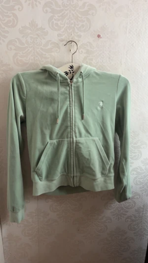 Mintgrön hoodie med dragkedja från Juicy Couture - Snygg mintgrön hoodie från Juicy Couture i mjuk velour med broderad logga på bröstet. Tröjan har huva med snörning, praktisk ficka framtill och hel dragkedja. Perfekt för chill eller när du vill vara extra comfy.