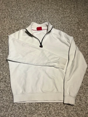 Vit half zip tröja från Hugo Boss - Vit långärmad tröja från Hugo Boss med svart half zip och ståkrage. Tröjan har ribbade muddar vid ärmslut och nederkant, samt en diskret röd detalj på insidan av kragen. (Har en liten, liten prick vid ärmen men syns inte vid användning) pris är diskuterbart