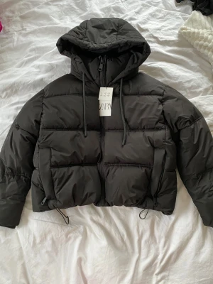 Svart pufferjacka med huva från Zara - Säljer en svart pufferjacka från Zara med croppad passform och stor huva. Jackan har snörning i nederkant, dragkedja framtill och två sidofickor med dragkedja. Perfekt inför vintern, så snygg!😍 Helt oanvänd och prislapp kvar🩷🩷🩷