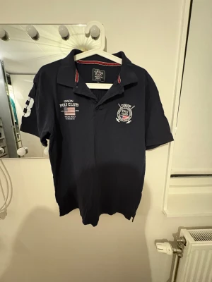 Mörkblå polo från Vinson Polo Club - Snygg mörkblå pikétröja från Vinson Polo Club med broderade detaljer och tryck på bröstet samt en stor vit siffra på ärmen. Klassisk krage med knappslå och korta ärmar. Materialet är mjuk bomull och insidan av kragen har rödvit randig kant.