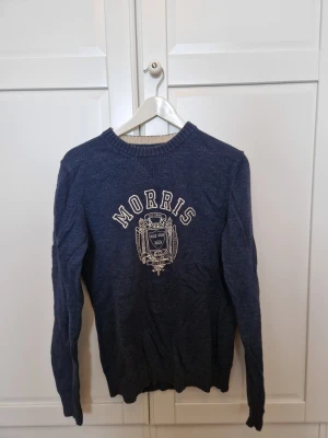 Mörkblå stickad tröja Morris Co - Snygg mörkblå stickad tröja från Morris Co i storlek medium. Tröjan har ett broderat emblem och text på bröstet, rund hals och ribbade muddar vid ärmslut och nederkant. Perfekt för dig som gillar klassisk stil med en modern touch.