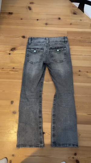 Grå bootcut jeans med låg midja - Säljer ett par grå jeans med snygg tvätt och låg midja. Modellen har bootcut-ben och klassiska fem fickor, med coola detaljer på bakfickorna. Jeansen är i stretchigt denim och har knappar och dragkedja framtill.