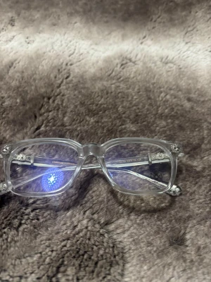 Transparanta glasögon från Chrome Hearts - Snygga rektangulära glasögon från Chrome Hearts med transparent båge och coola silverdetaljer vid gångjärnen. Tillverkade i Italien och har en unik vibe med små metallblommor på sidorna. Perfekt för dig som vill sticka ut med stilrena men ändå lyxiga glasögon.