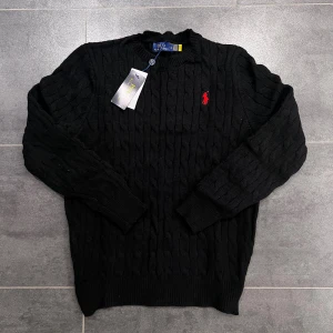 Svart Polo Ralph Lauren - Svart kabelstickad tröja från Polo Ralph Lauren med klassisk rund hals och röd broderad logga på bröstet. Tröjan har långa ärmar och ribbade muddar vid ärmslut och nederkant. Perfekt för dig som gillar stilren och tidlös design.