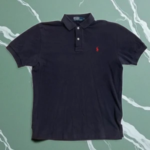 Mörkblå piké från Polo Ralph Lauren - Klassisk mörkblå pikétröja från Polo Ralph Lauren med röd broderad logga på bröstet. Tröjan har korta ärmar, krage och knäppning med två vita knappar. Tillverkad i bomull för en skön och luftig känsla.