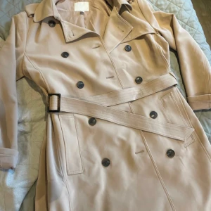 Beige trenchcoat från H&M, strl 42 - Snygg beige trenchcoat från H&M i storlek 42. Jackan har klassisk dubbelknäppning, bälte i midjan med spänne och långa ärmar. Materialet är mjukt och slätt, perfekt för vår och höst. Stilren design med svarta knappar och krage.