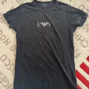 Svart t-shirt från Emporio Armani - Svart t-shirt från Emporio Armani med klassisk rund hals och korta ärmar. Framtill finns en vit EA-logga med den ikoniska örnen. T-shirten är i mjuk bomull och har en clean, stilren look som passar perfekt till jeans eller shorts.