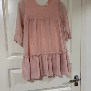 Ljusrosa volangklänning med prickar - Supersöt ljusrosa kortklänning med volangkant och diskret prickigt mönster. Klänningen har trekvartsärm och är tillverkad i ett tunt, lätt material som ger ett luftigt intryck. Perfekt för vår och sommar när du vill känna dig extra fin.