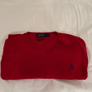 Röd v-ringad tröja Polo Ralph Lauren - Riktigt snygg röd v-ringad tröja från Polo Ralph Lauren i storlek L. Tröjan är gjord av mjuk pima bomull och har långa ärmar samt den klassiska blå Polo-loggan broderad på bröstet. Perfekt för dig som gillar stilrena och tidlösa plagg.