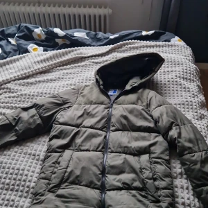 Olivgrön pufferjacka Jack & Jones - Säljer en olivgrön pufferjacka från Jack & Jones med huva och hel dragkedja framtill. Jackan har quiltad design och är tillverkad i syntetmaterial, perfekt för kyliga dagar. Enkel och stilren look som funkar till de flesta outfits.