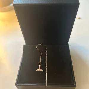 Silvriga örhängen med valfena - Unika silvriga örhängen med tunn kedja och en liten valfena som hänge. Snygg minimalistisk design som ger en cool och modern vibe. Perfekt för dig som gillar havsinspirerade detaljer och stilrena smycken.