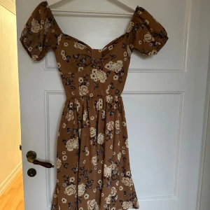 Brun blommig kortklänning med snörning - Supersöt brun kortklänning med blommigt mönster och puffärmar. Klänningen har offshoulder-modell, snörning och öppen rygg som ger en unik look. Perfekt för sommarens alla tillfällen. Materialet känns lätt och flowy.