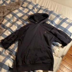 Svart hoodie från GANT med huva - Mörk blå Hoodie från GANT i storlek M med klassisk huva och känguruficka framtill. Tröjan har GANT-logga broderad på bröstet och ribbade muddar vid ärmslut. Perfekt för chill dagar och enkel att matcha med jeans eller joggers.