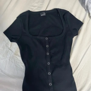 Svart  topp från Gina Tricot - Svart ribbad topp från Gina Tricot i storlek XS. Modellen har korta ärmar, djup fyrkantig halsringning och knappar framtill. Materialet är mjukt och stretchigt, perfekt för en chill och stilren look. Snygg att matcha med jeans eller kjol.