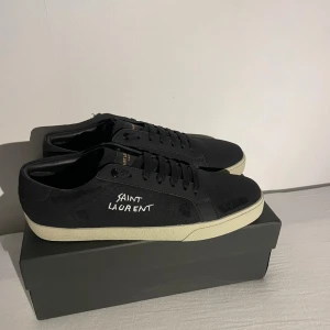 Svarta Saint Laurent sneakers - Svarta sneakers från Saint Laurent med vit broderad logga på sidan och gulaktig platt sula. Skorna har snörning, rund tå och är tillverkade i canvas och skinn. Klassisk och stilren design med Saint Laurent Paris-detaljer på plös och sula.