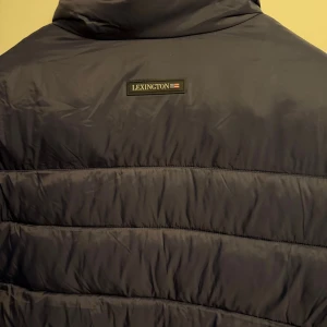 Svart pufferjacka från Lexington - Säljer en svart pufferjacka från Lexington med beige insida. Jackan har ståkrage, två sidofickor med dragkedja och quiltad design. Perfekt för dig som vill ha en clean och stilren look under höst och vinter. Dragkedja framtill för enkel på- och avtagning.