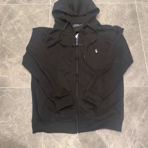 Svart hoodie från Polo Ralph Lauren - Svart hoodie från Polo Ralph Lauren med dragkedja och huva. Klassisk design med vit broderad logga på bröstet, snörning i huvan och ribbade muddar. Perfekt för chill dagar och enkel att matcha med jeans eller joggers.
