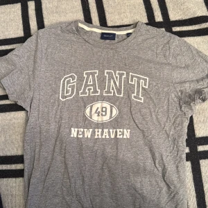Grå GANT t-shirt med tryck - Snygg grå t-shirt från GANT med klassiskt New Haven-tryck och siffran 49 framtill. T-shirten har rund hals och korta ärmar, tillverkad i mjuk bomull för skön känsla. Perfekt för dig som gillar sportig och avslappnad stil.